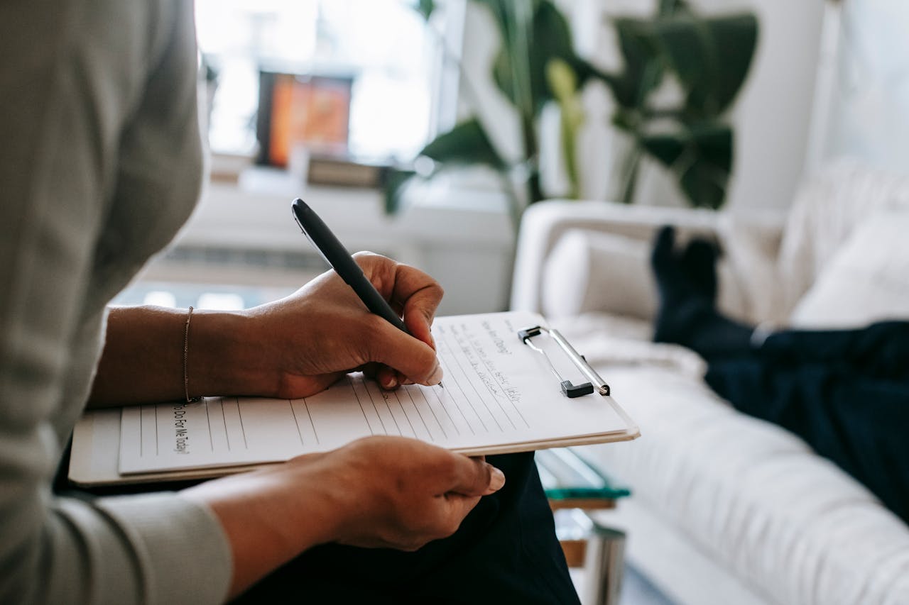 creative Unrecognizable ethnic female therapist taking notes on clipboard while filling out form during psychological appointment with anonymous client lying on blurred background